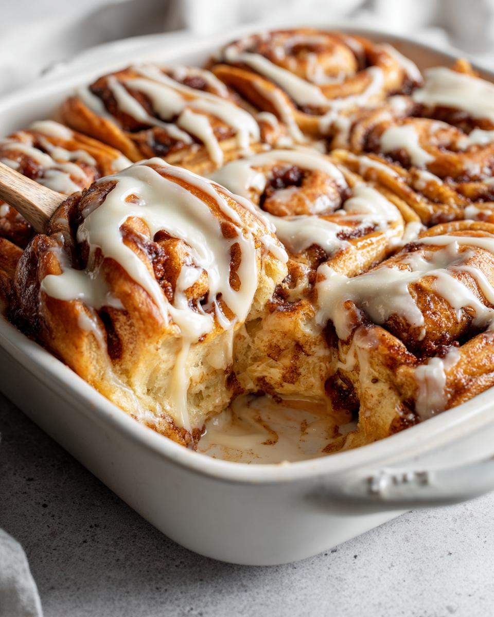 Close-up of a freshly baked cinnamon roll casserole with creamy icing being lifted with a wooden spoon.