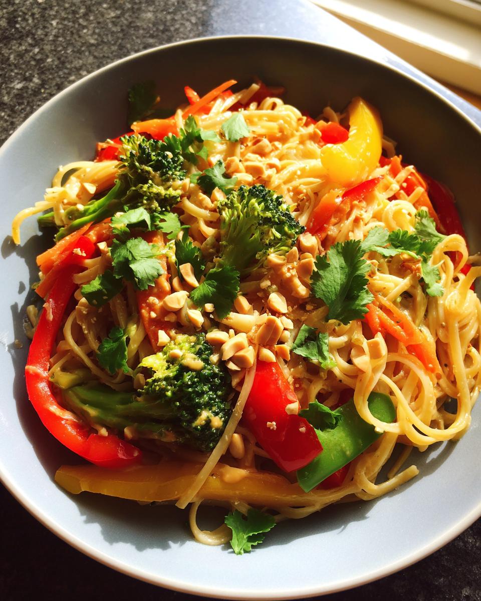A vibrant bowl of Thai peanut noodles with vegetables, including broccoli, peppers, and carrots, topped with peanuts and cilantro.