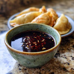 Close-up of a bowl of dumpling sauce with sesame seeds and chili flakes, with dumplings in the background.