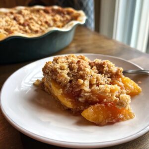 A slice of Peach Crisp with Fresh Peaches on a white plate, showcasing the juicy peaches and golden-brown crisp topping.