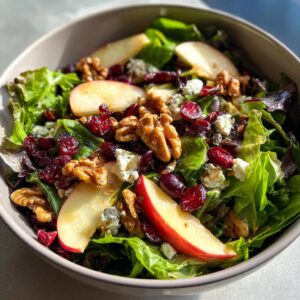 A close-up view of a vibrant apple walnut salad, featuring crisp lettuce, apple slices, walnuts, dried cranberries, and crumbled blue cheese.