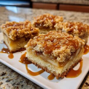 Close-up of four caramel apple pie bars on a white plate, drizzled with caramel sauce.