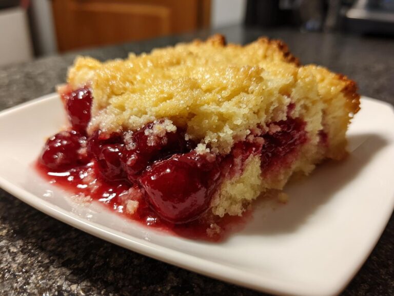 A close-up of a slice of Cherry Dump Cake on a white plate, showcasing juicy cherries and a golden cake topping.