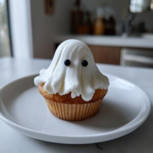 A single adorable ghost cupcake with white frosting and black candy eyes on a white plate.