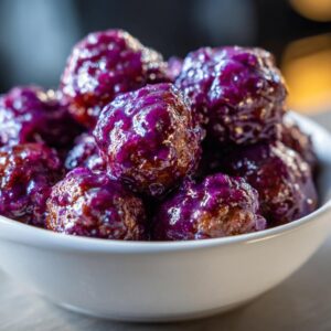 A close-up of a white bowl filled with glistening grape jelly meatballs.