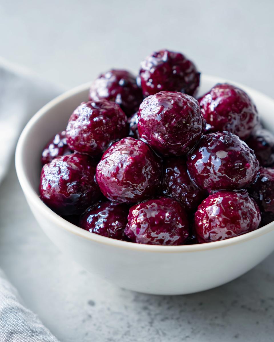 A close-up of glossy grape jelly meatballs piled high in a white bowl.