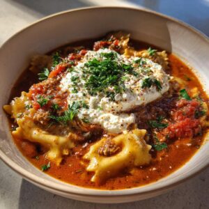 A close-up of a bowl of hearty Lasagna Soup, featuring ravioli, rich tomato broth, and a dollop of creamy ricotta topped with fresh parsley.