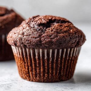 A close-up view of a moist chocolate cupcake in a paper liner, with another cupcake blurred in the background.