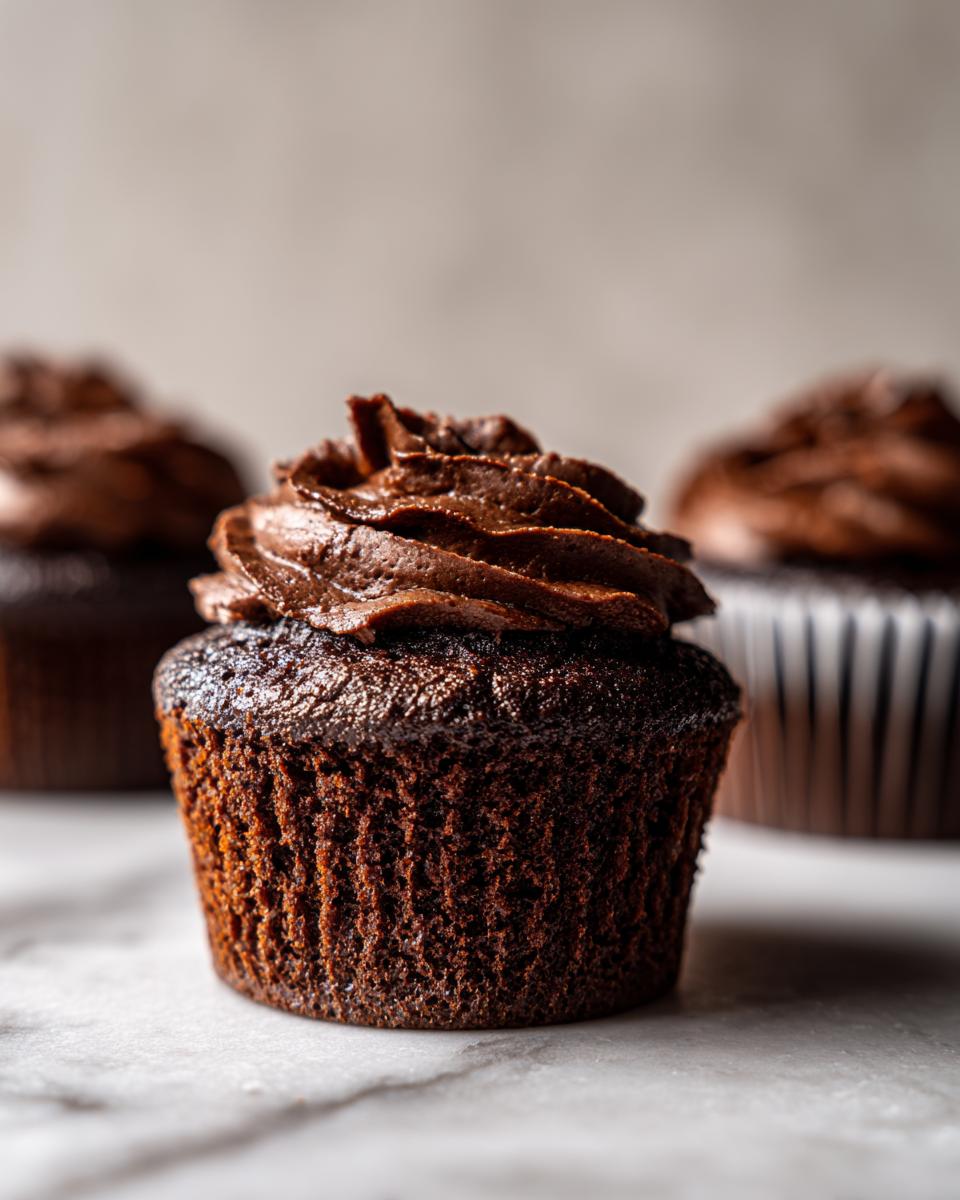 Close-up of a moist chocolate cupcake with rich chocolate frosting, with two more chocolate cupcakes blurred in the background.