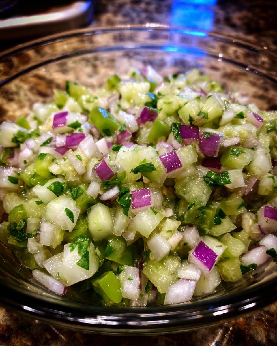 A close-up of a glass bowl filled with fresh Pickle de Gallo, featuring diced cucumber, red onion, green pepper, and cilantro.