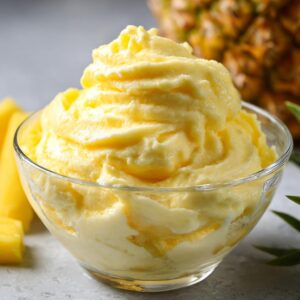 Close-up of a creamy Pineapple Dole Whip copycat in a glass bowl, with pineapple chunks and a whole pineapple in the background.