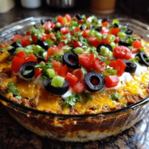A close-up of a freshly made Seven Layer Dip in a glass dish, topped with tomatoes, olives, and green onions.