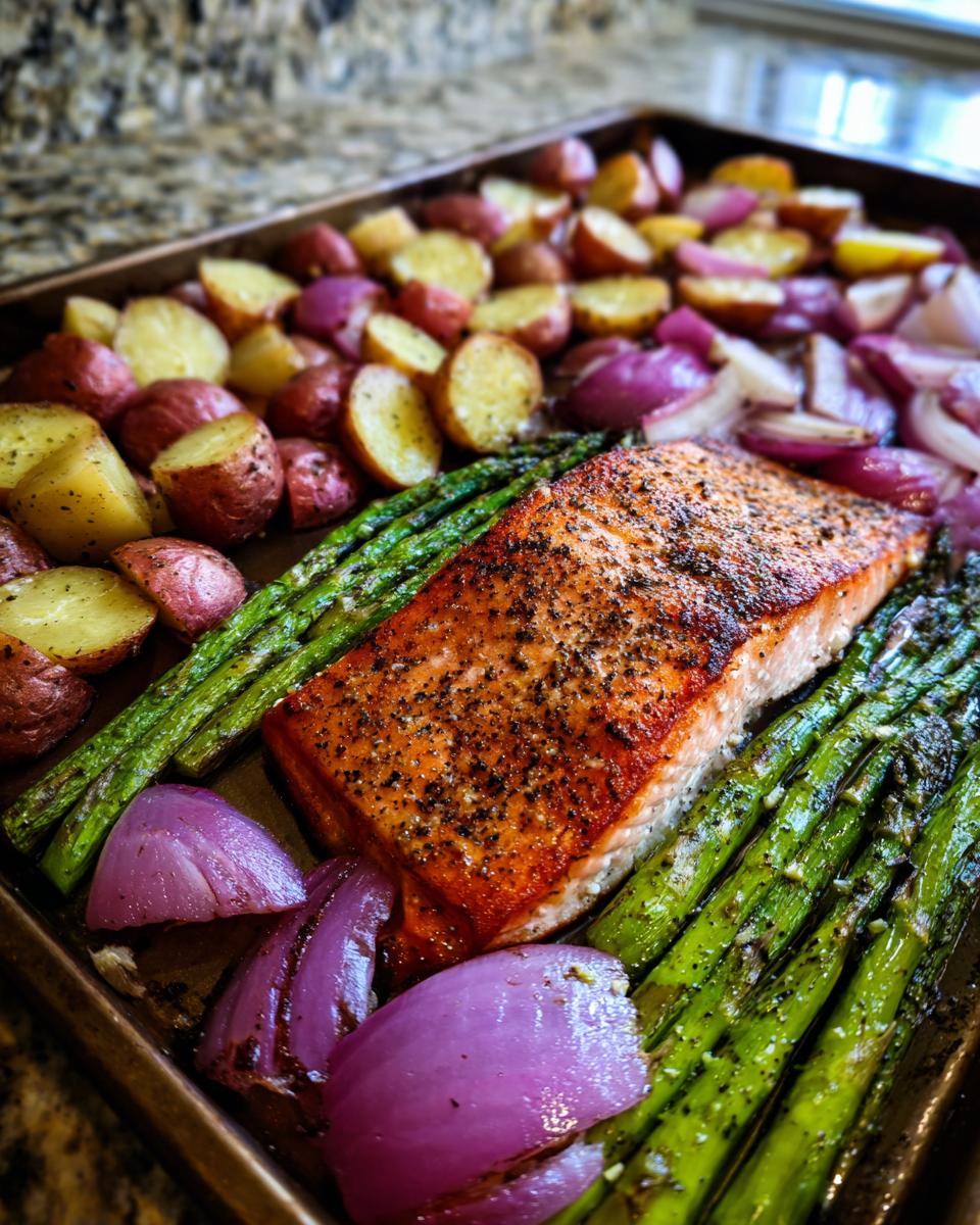A close-up of a sheet pan filled with salmon, asparagus, red onion wedges, and halved baby potatoes.