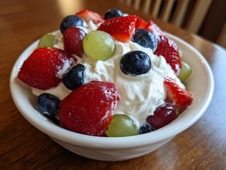 Close-up of a white bowl filled with creamy topping, strawberries, blueberries, and grapes for a cheesecake fruit salad.