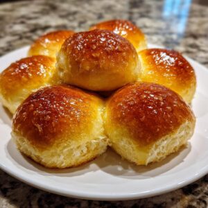 A stack of six golden brown, glossy dinner rolls with a rich buttery glaze on a white plate.