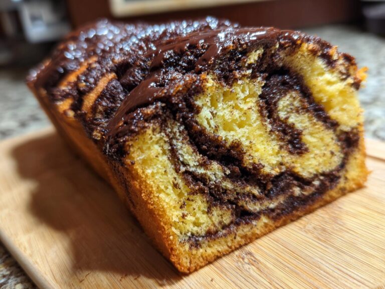 A close-up of a freshly baked chocolate babka loaf, showing the beautiful yellow and dark chocolate swirl pattern inside and a shiny chocolate glaze on top.