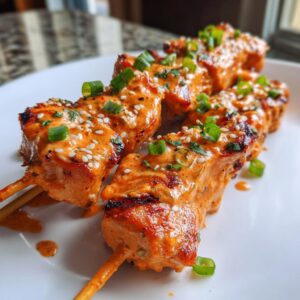 Close-up of grilled bang bang chicken skewers generously coated in orange sauce, topped with sesame seeds and green onions.