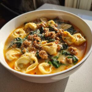A close-up of a steaming white bowl filled with creamy sausage tortellini soup, featuring spinach and parmesan.