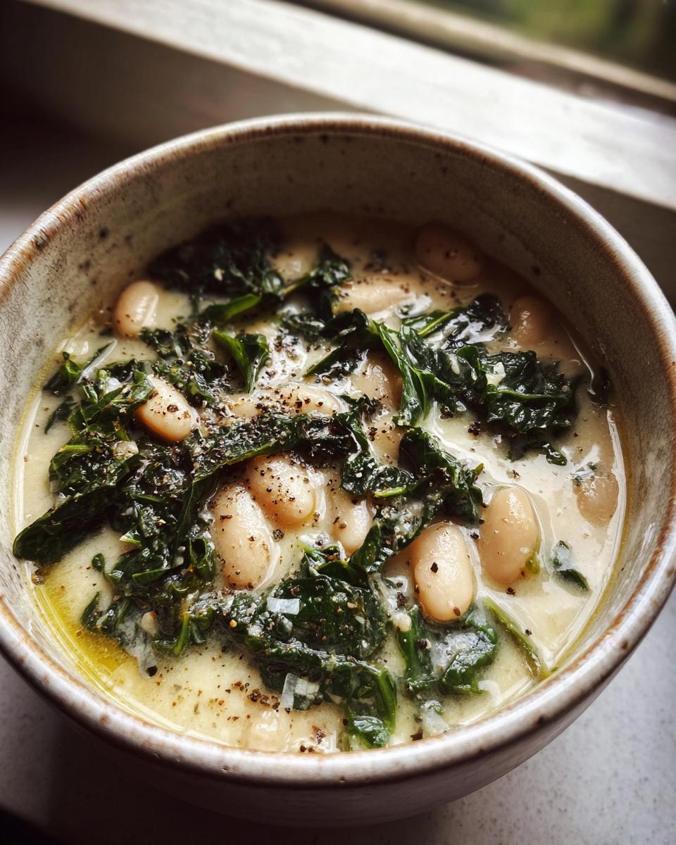 Close-up of creamy tuscan white bean soup featuring cannellini beans and dark green kale, topped with cracked black pepper.