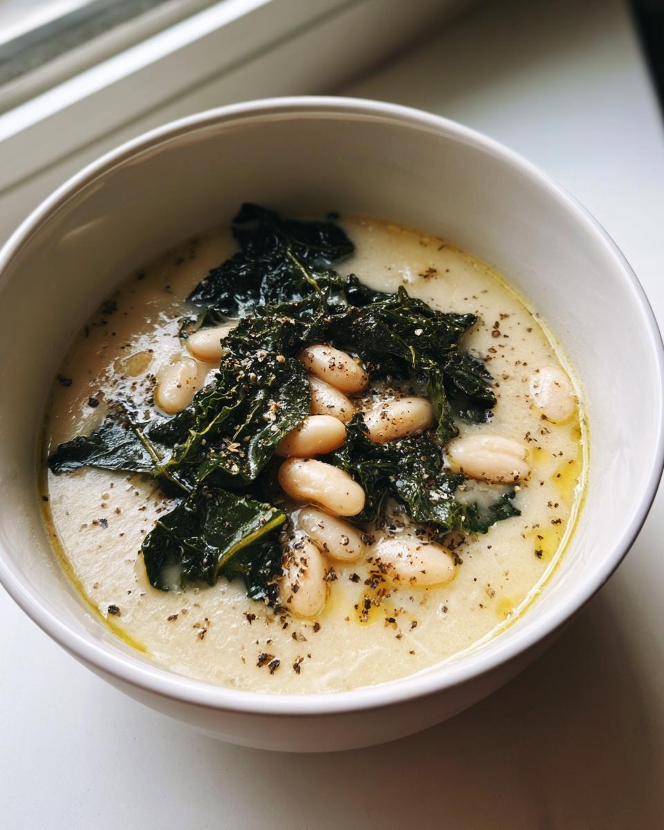 A close-up of creamy tuscan white bean soup topped with whole white beans, dark green kale, and cracked black pepper.
