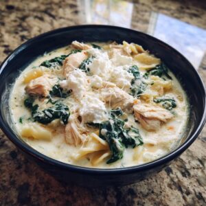 A close-up of a dark bowl filled with creamy white chicken lasagna soup, topped with shredded chicken, spinach, and dollops of ricotta cheese.