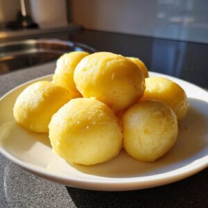A close-up of several fluffy, golden potato dumplings piled on a white plate in a sunlit kitchen.