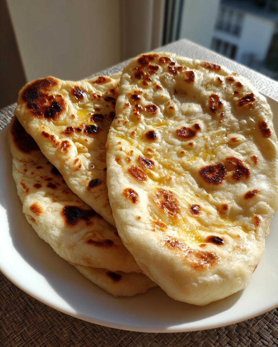 Three pieces of soft, freshly made homemade naan bread stacked on a white plate, showing charred spots and melted butter.