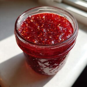 Close-up of vibrant, homemade raspberry jam, showing seeds, sealed in a quilted glass jar near a window.