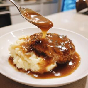 A spoonful of rich brown chicken gravy is being poured over a piece of meat resting on mashed potatoes.