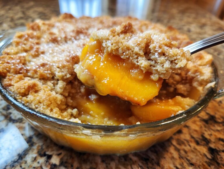 A spoonful of warm, syrupy peaches and crumbly topping being lifted from a glass dish of Southern Peach cobbler.