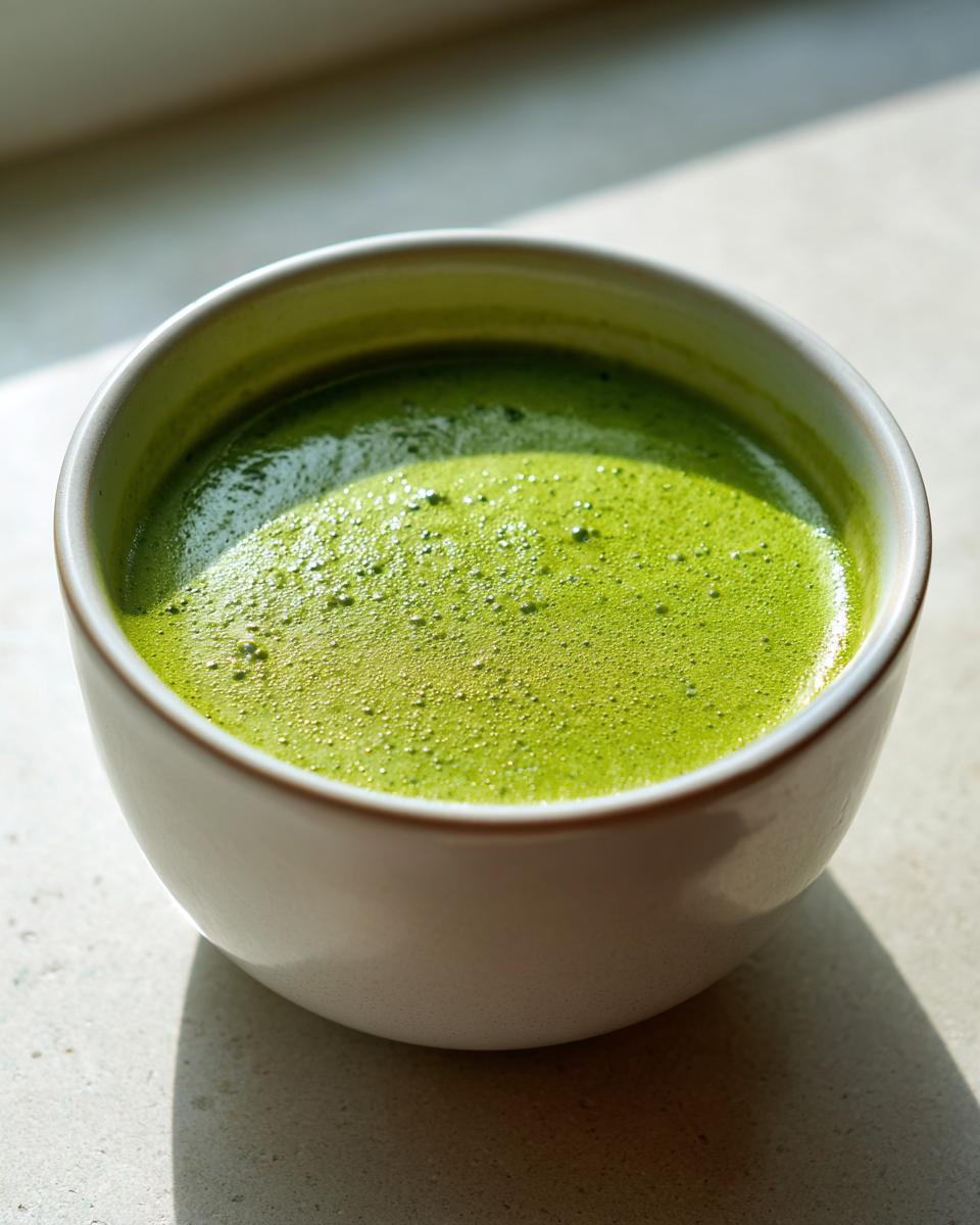 Close-up of vibrant green, frothy asparagus soup served in a small white bowl, highlighted by natural light.