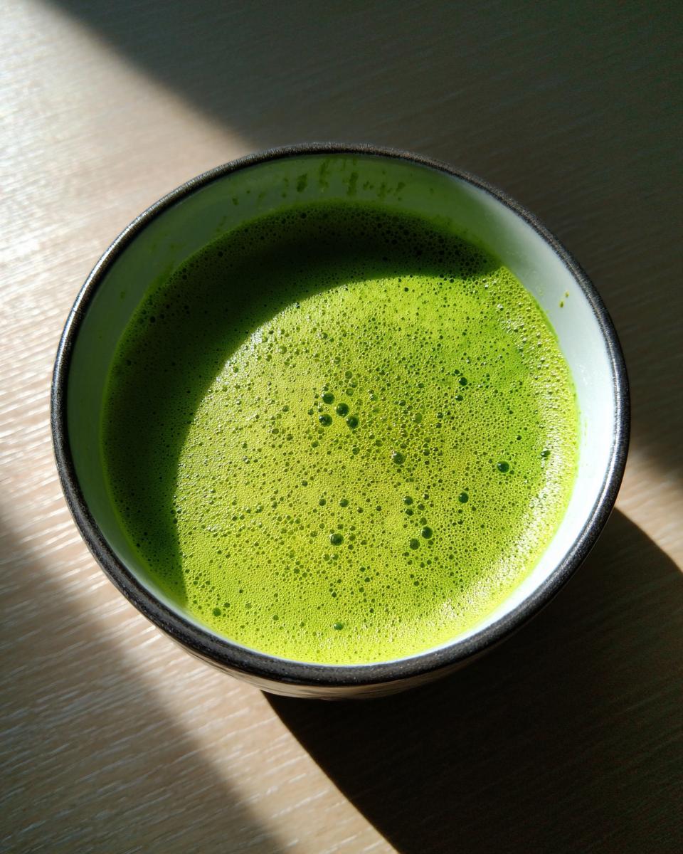 Overhead shot of a vibrant green bowl of creamy spinach soup with a frothy top, illuminated by sunlight.
