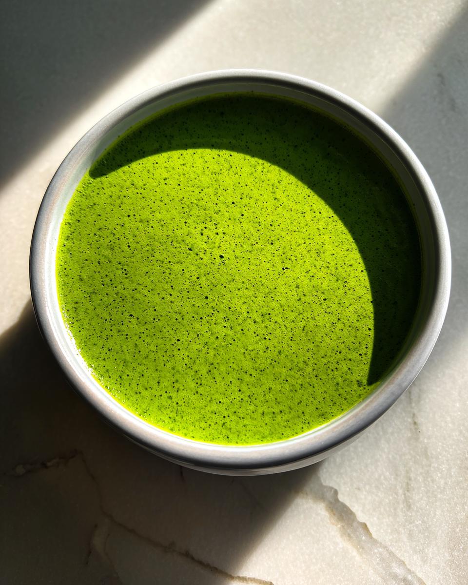 Overhead view of a bowl of bright green spinach soup with a frothy top, set against a marble surface.