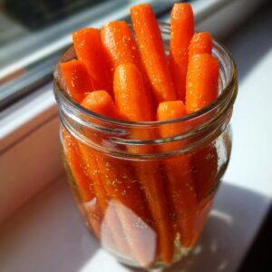Bright orange, whole pickled carrots packed tightly in a clear glass jar, seasoned with visible herbs.