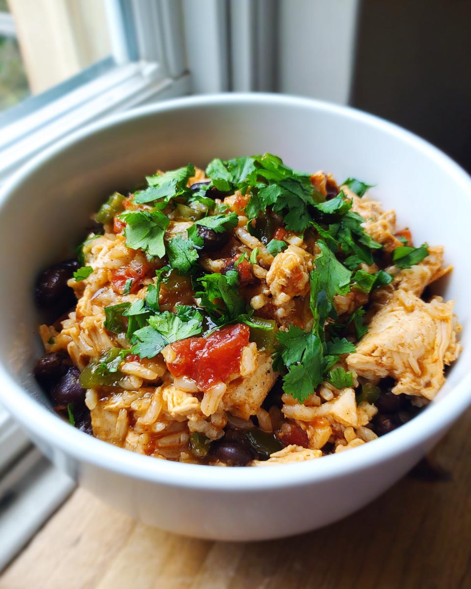 A white bowl filled with a colorful chicken burrito bowl mixture of rice, shredded chicken, black beans, tomatoes, and topped with fresh cilantro.