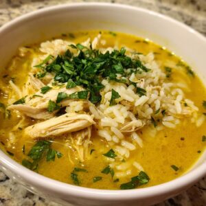 Close-up of a bowl of Lemon Ginger Turmeric Chicken and Rice Soup, topped with shredded chicken, white rice, and fresh parsley.