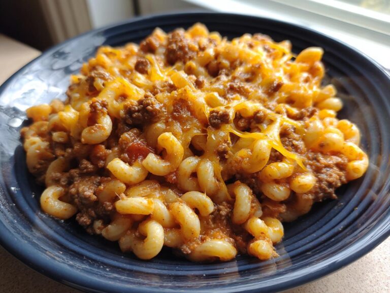 A close-up of a dark blue bowl filled with cheesy taco pasta featuring elbow macaroni, seasoned ground beef, and melted cheddar cheese.