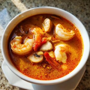 Close-up of a steaming white bowl filled with vibrant orange and red tom yum soup, featuring large shrimp and sliced mushrooms.