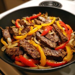 Close-up of sizzling steak fajita strips with red and yellow peppers and onions in a black skillet.
