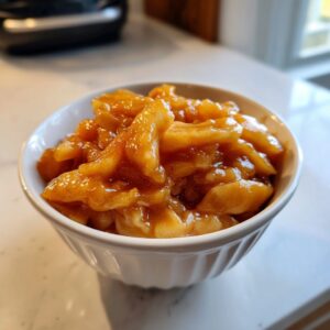 A white bowl filled with glossy, spiced apple pie filling, showcasing thick slices of apple coated in cinnamon sauce.