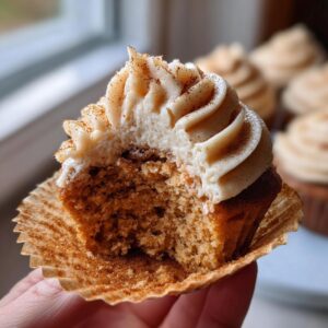A hand holds a bitten churro cupcake showing the moist interior and cinnamon-dusted frosting.