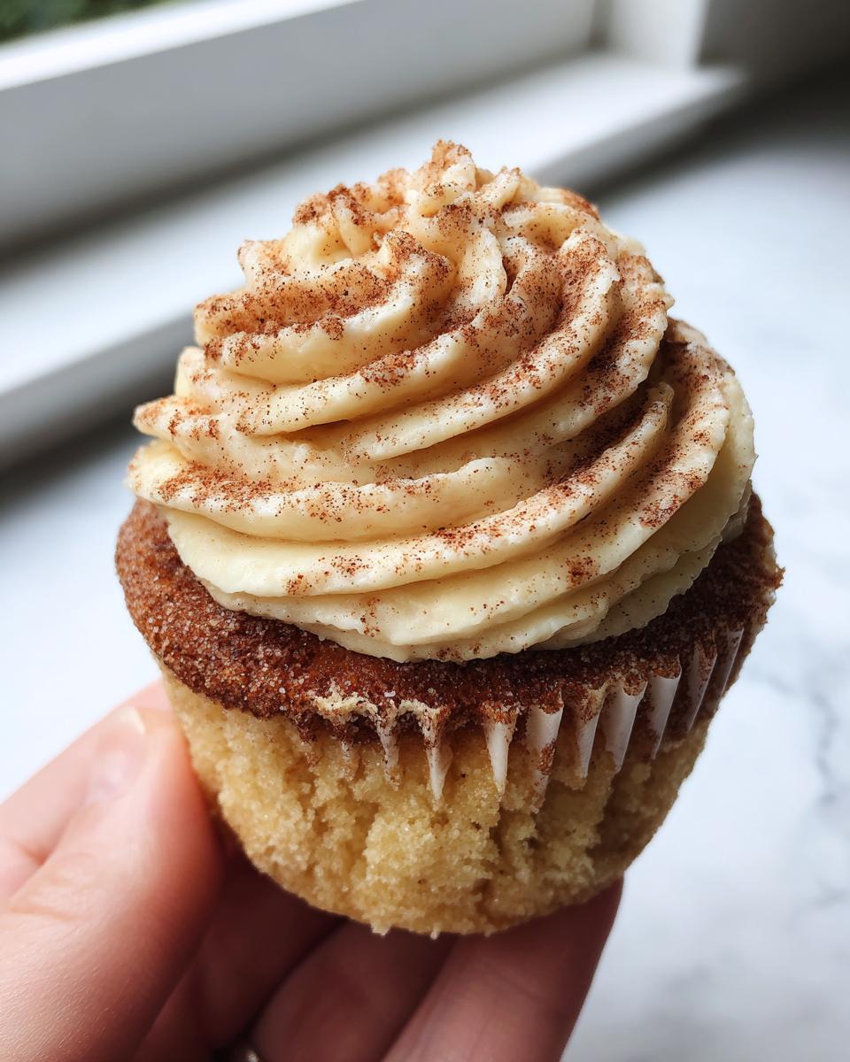 A close-up shot of a hand holding a perfectly frosted churro cupcake dusted with cinnamon sugar.