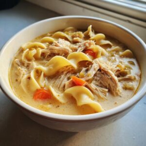 Close-up of a white bowl filled with creamy chicken noodle soup featuring shredded chicken, wide egg noodles, and carrots.