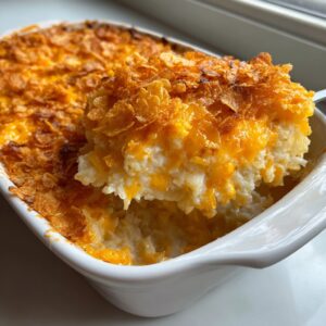 A spoonful of creamy hashbrown casserole topped with melted cheese and crispy cornflakes being lifted from a white baking dish.