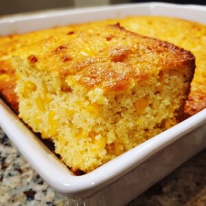 A close-up of a golden, moist slice of jiffy corn casserole being lifted from a white baking dish.