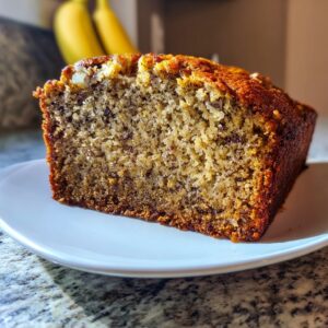 Close-up of a moist slice of homemade banana bread recipe on a white plate, with whole bananas blurred in the background.