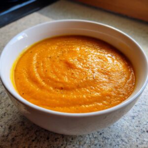 Close-up of a vibrant orange bowl of creamy carrot ginger soup, lightly seasoned with black pepper.