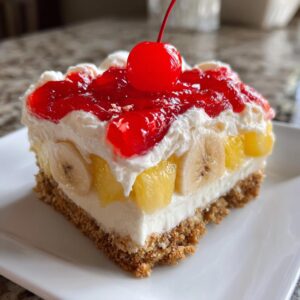 A perfect square slice of banana split cake showing layers of graham cracker crust, cream filling, bananas, pineapple, and topped with whipped cream and a cherry.