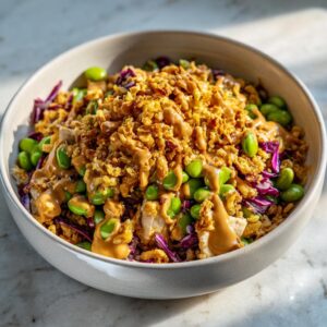 A bowl of crispy rice salad topped with edamame, red cabbage, crispy fried onions, and drizzled with peanut sauce.
