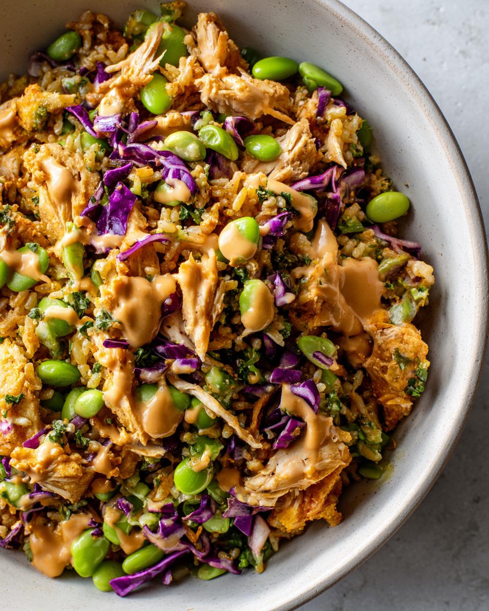 Overhead close-up of a bowl filled with crispy rice salad, shredded chicken, bright green edamame, purple cabbage, and drizzled with peanut sauce.